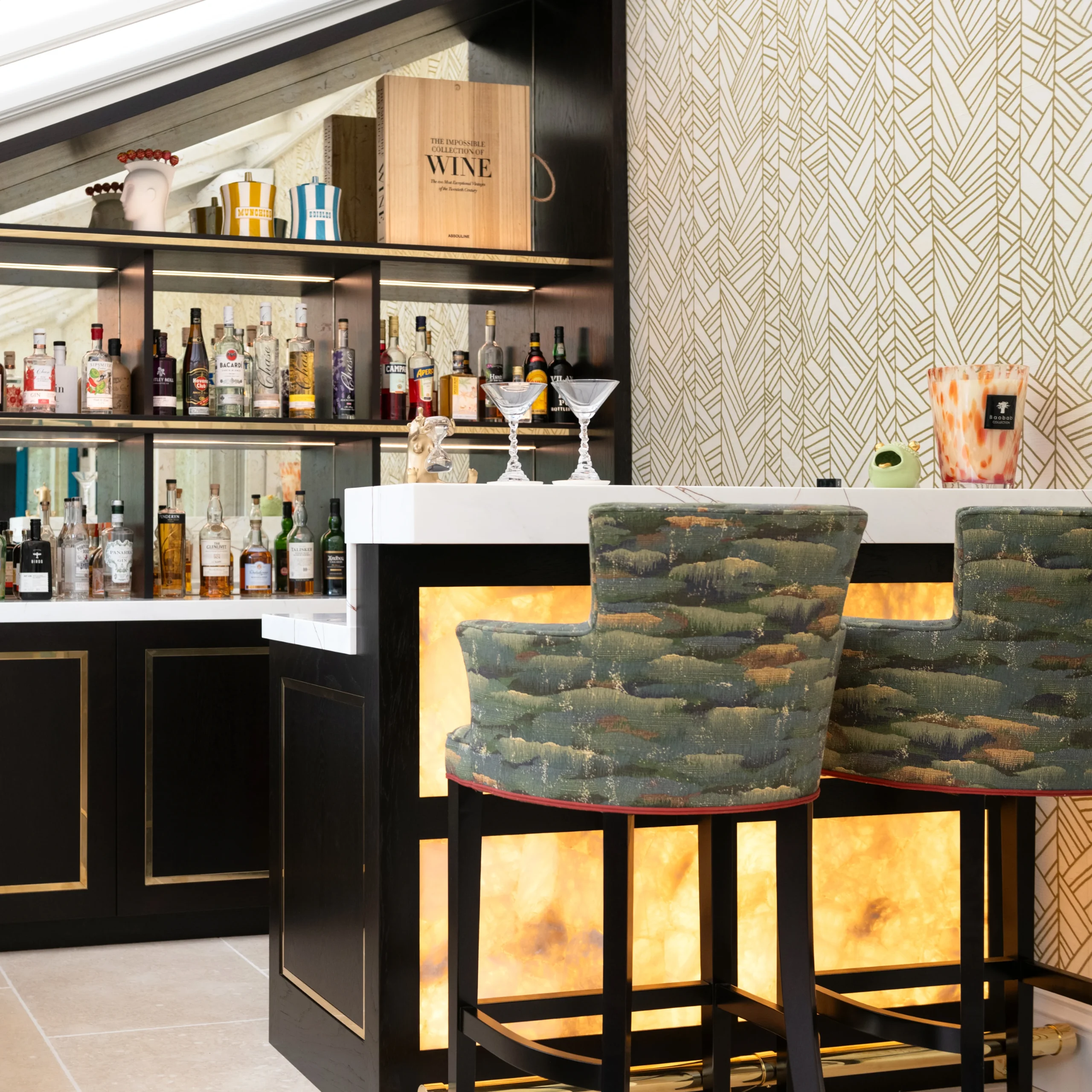 An atmospheric Tobias Oliver home bar with stocked shelving, patterned stools and wallpapered walls